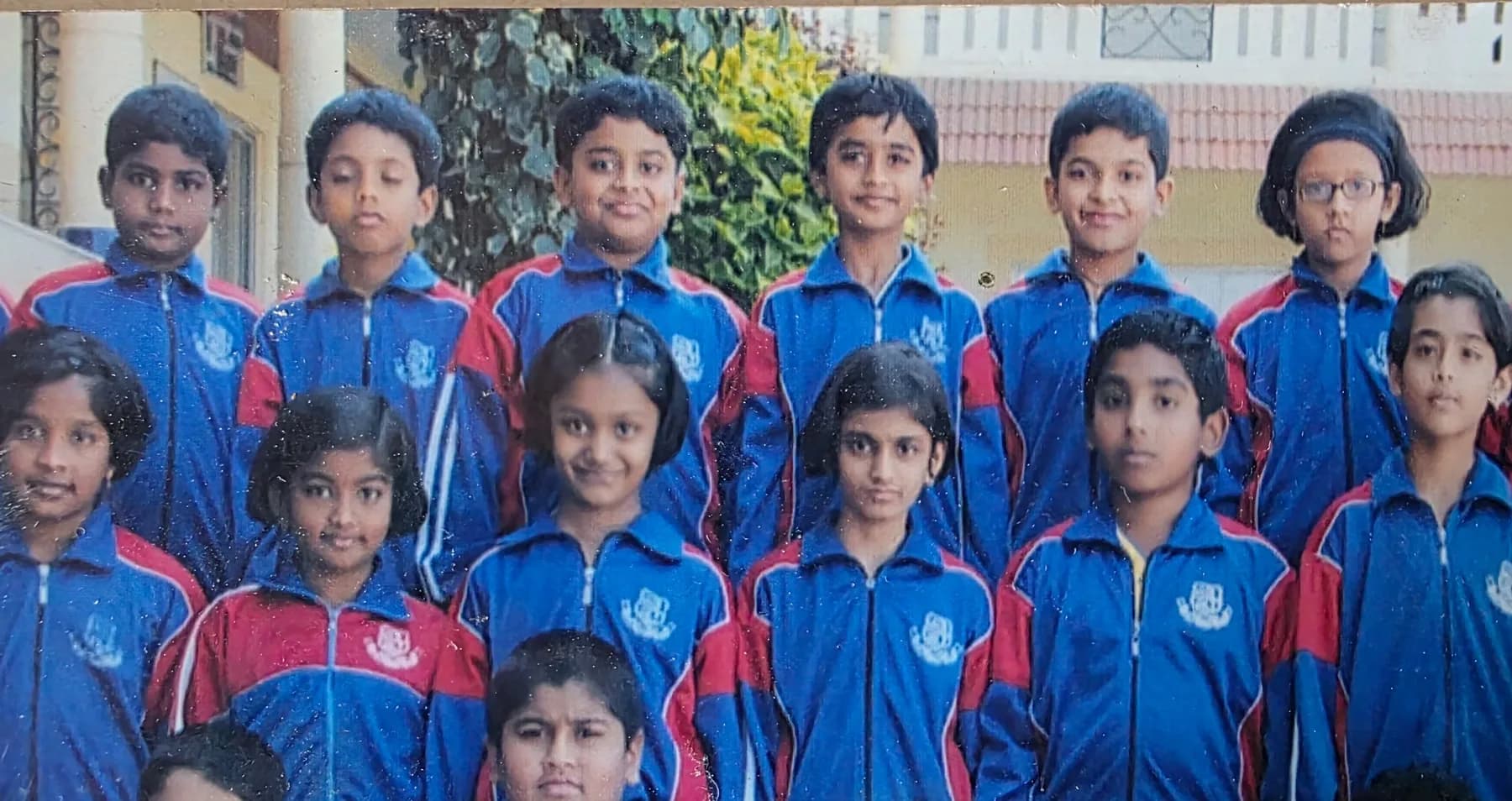 A class photograph from 2006. Fifteen children in blue and red school tracksuits stand in rows outside the school building. Two boys in the back row, Samarth and Dhanush, are circled.